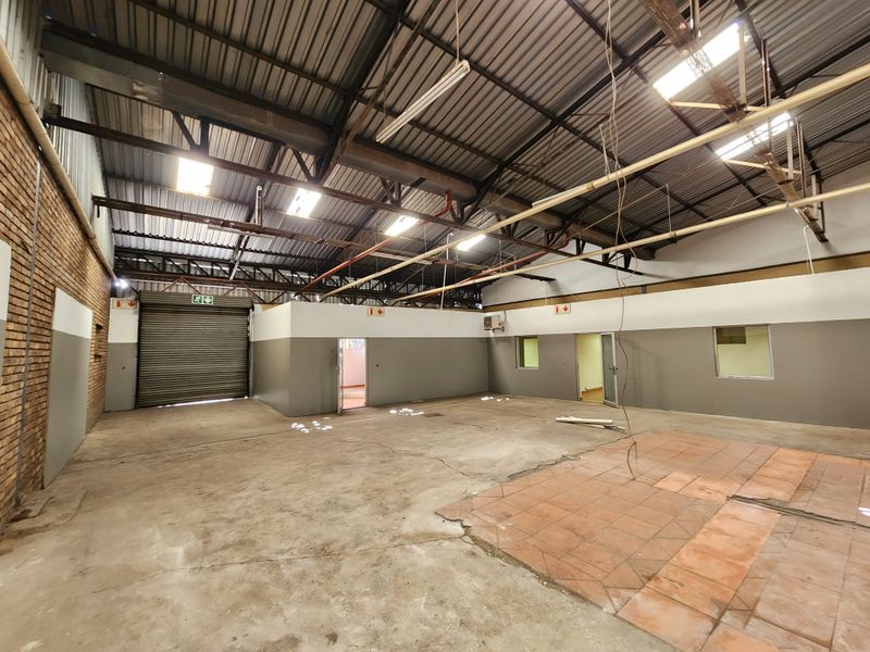 Spacious industrial utility area featuring high ceilings with exposed steel trusses, roller shutter door access, and a combination of concrete and tiled flooring.