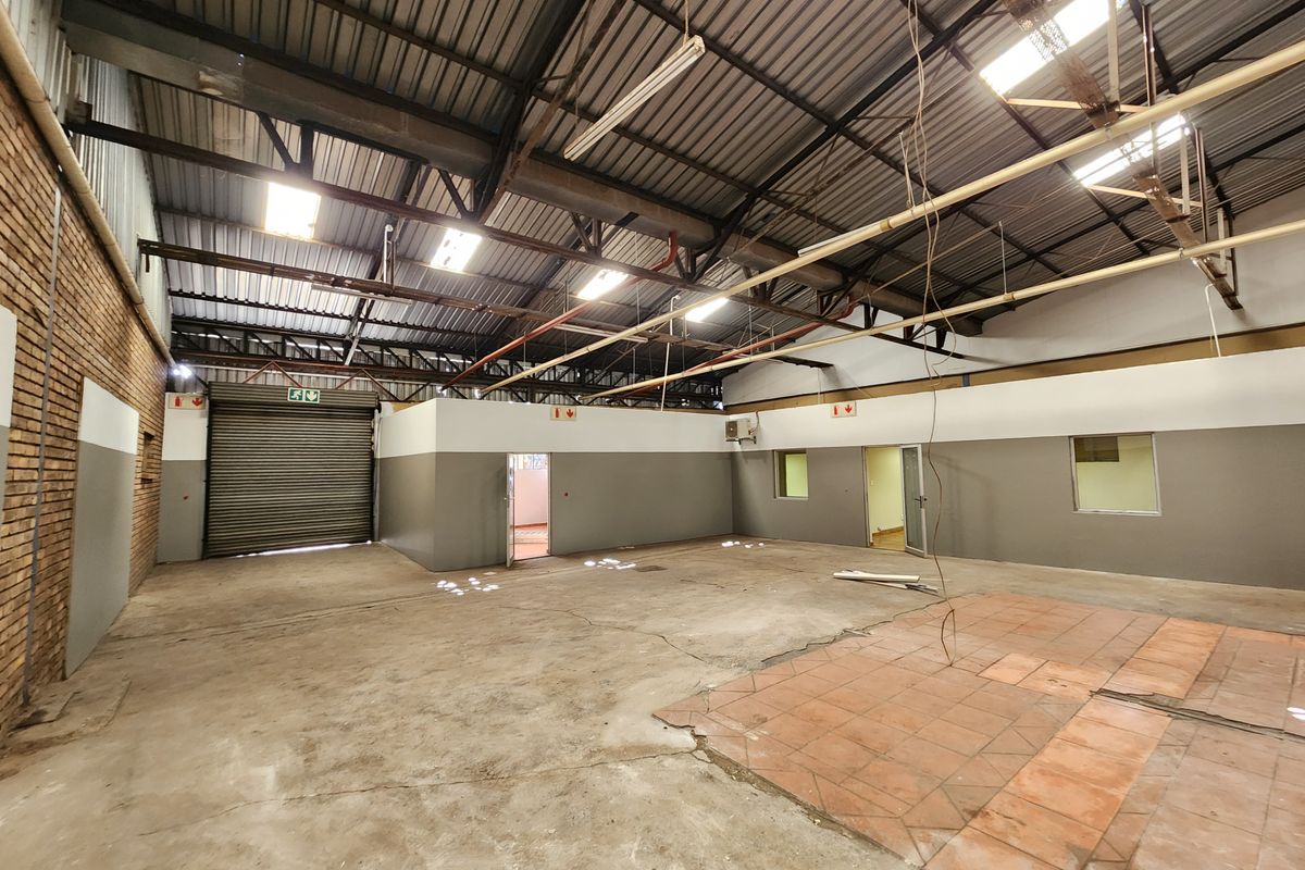 Spacious industrial utility area featuring high ceilings with exposed steel trusses, roller shutter door access, and a combination of concrete and tiled flooring.