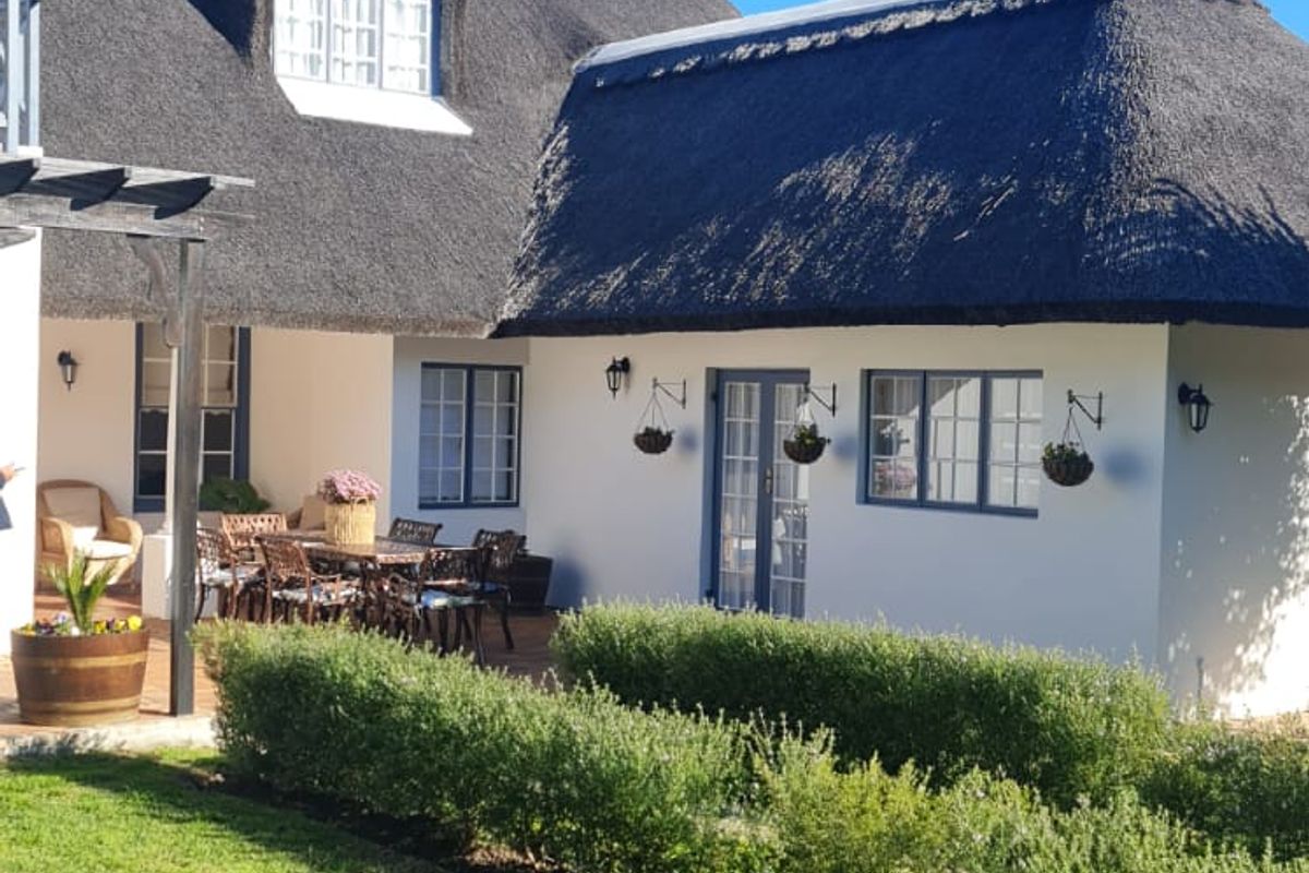 A well-maintained rear exterior view of a guest house featuring a traditional thatched roof, a paved patio area, and a neatly landscaped garden.