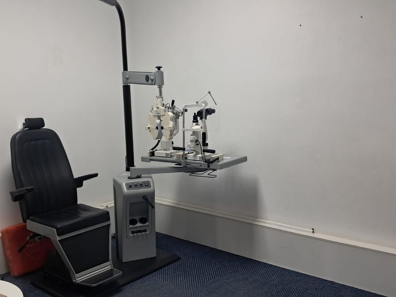 A functional clinical examination room featuring carpeted flooring and standard wall finishes, currently equipped with specialized optometry diagnostic equipment.
