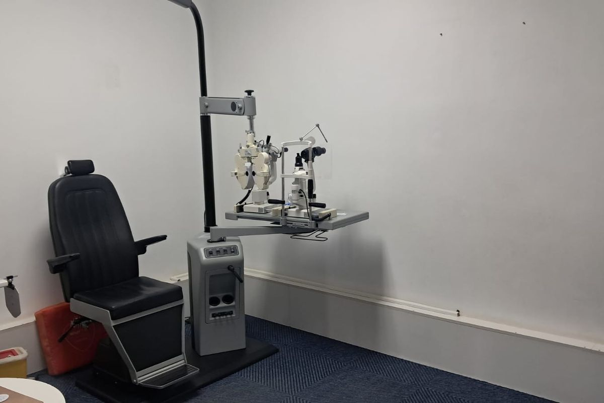 A functional clinical examination room featuring carpeted flooring and standard wall finishes, currently equipped with specialized optometry diagnostic equipment.