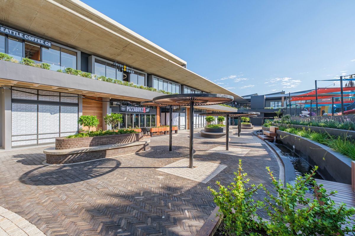 A modern outdoor commercial plaza featuring paved walkways, built-in brick planters, water features, and shaded seating areas in front of restaurant storefronts.