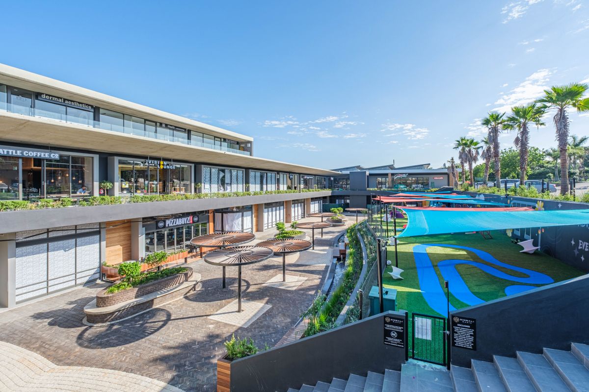 Modern commercial complex featuring a multi-level retail plaza with paved outdoor seating areas, integrated landscaping, and a vibrant recreational play zone.
