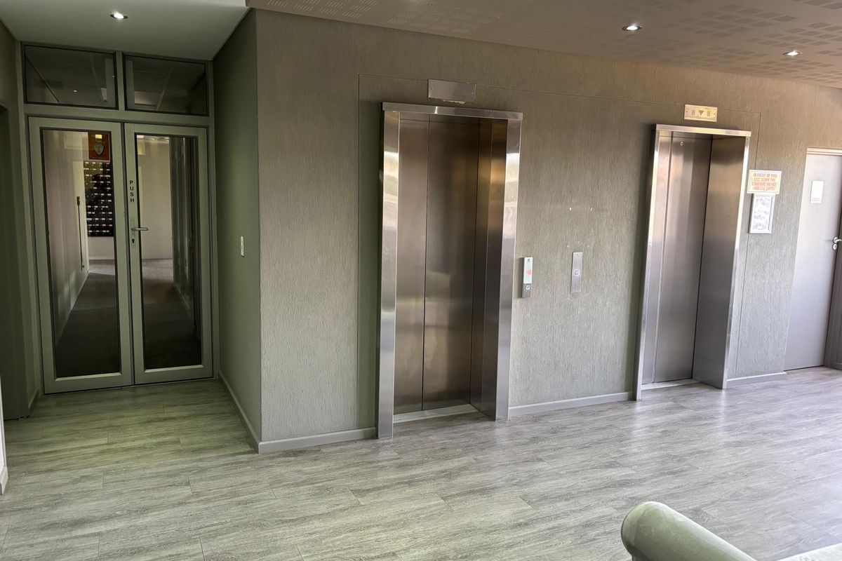 Modern office building lobby featuring dual stainless steel elevators, glass entrance doors, and contemporary laminate flooring with recessed ceiling lighting.