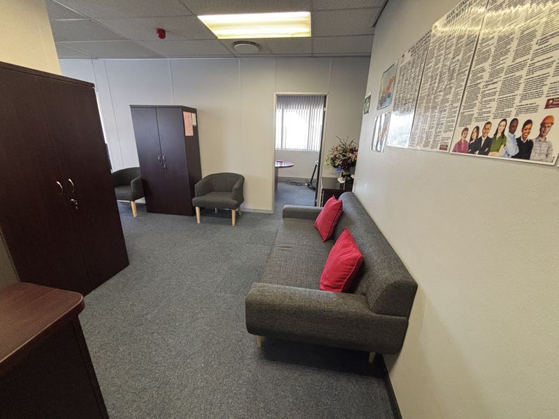 A functional office waiting area featuring carpeted flooring, a suspended ceiling with recessed lighting, and an adjoining room visible through the doorway.
