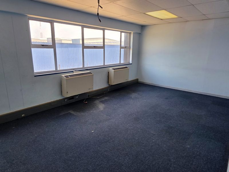 An empty office space featuring large windows, wall-mounted air conditioning units, suspended ceiling tiles, and dark carpet flooring, suitable for commercial use.