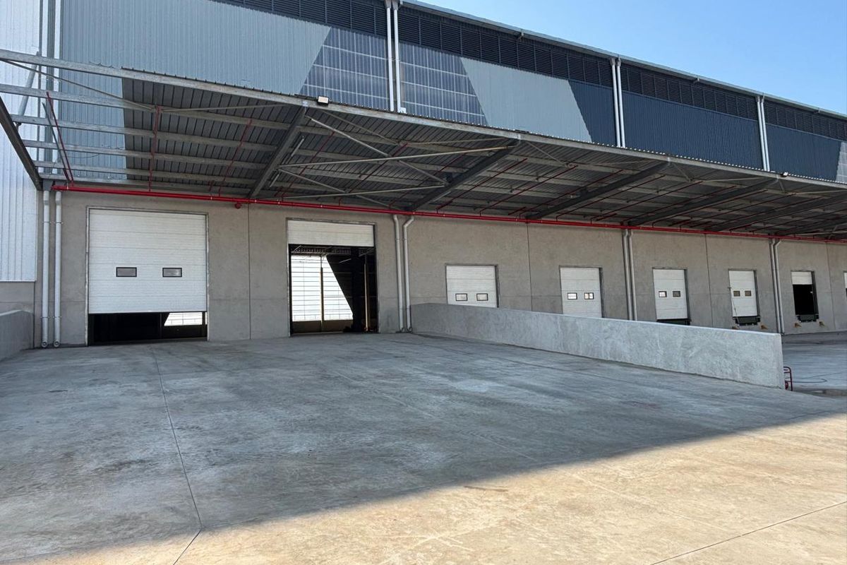 Industrial warehouse exterior featuring multiple roller shutter loading doors, a concrete loading bay, and a large steel-framed canopy structure.