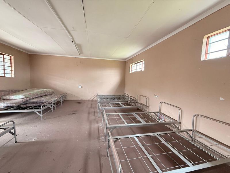 A large, utilitarian industrial room featuring multiple metal bed frames, high ceilings, and small security-style windows.