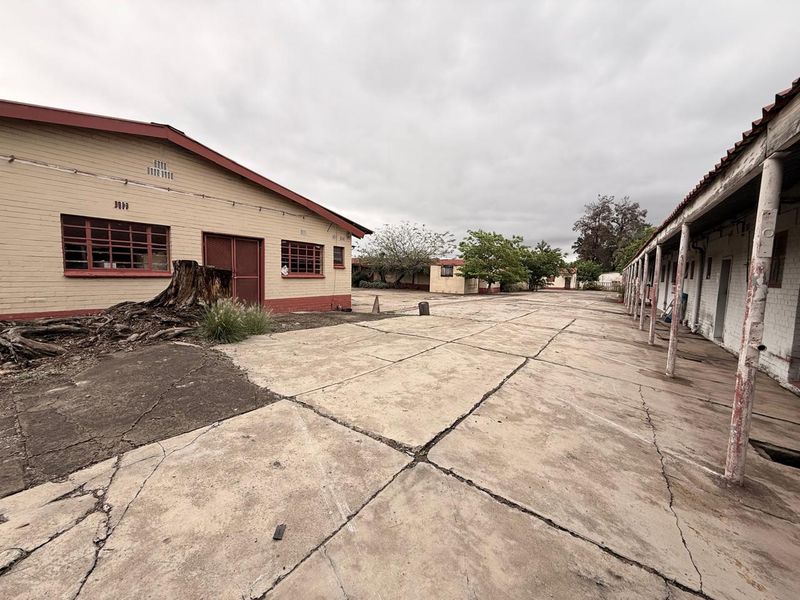 Expansive industrial courtyard featuring weathered concrete paving, covered walkways with structural columns, and multiple single-story brick buildings.