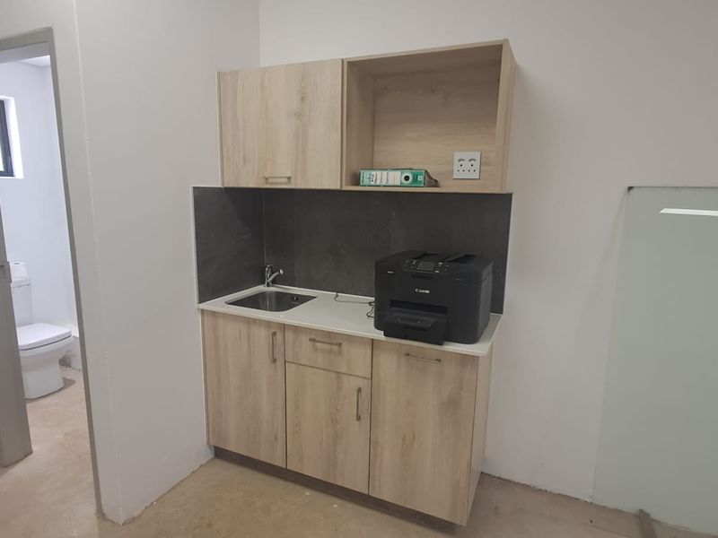 A compact kitchenette featuring light wood-finish cabinetry, a stainless steel sink, and a dark stone-look backsplash, adjacent to a small restroom.