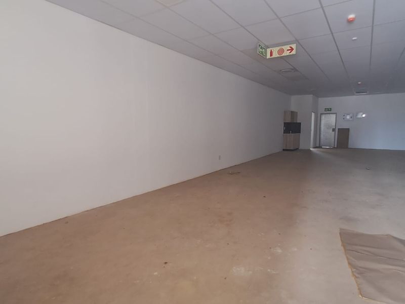A spacious, vacant open-plan retail unit featuring a suspended ceiling, recessed lighting, and a small built-in kitchenette area.