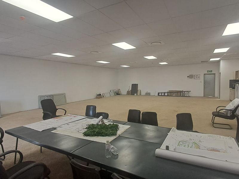 A large, open-plan commercial space featuring a suspended ceiling with recessed lighting, carpet flooring, and a rear fire exit door.