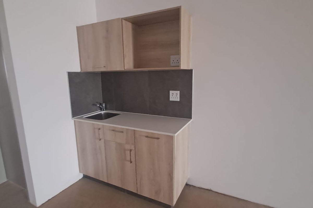 A compact kitchenette featuring light wood-finish cabinetry, a stainless steel sink, and a modern tiled splashback in a clean, minimalist space.