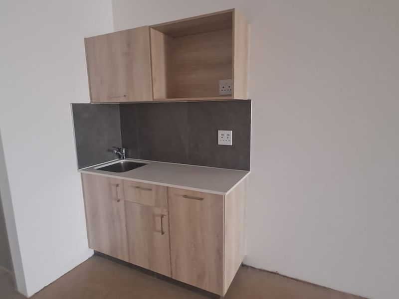 A compact kitchenette featuring light wood-finish cabinetry, a stainless steel sink, and a modern tiled splashback in a clean, minimalist space.