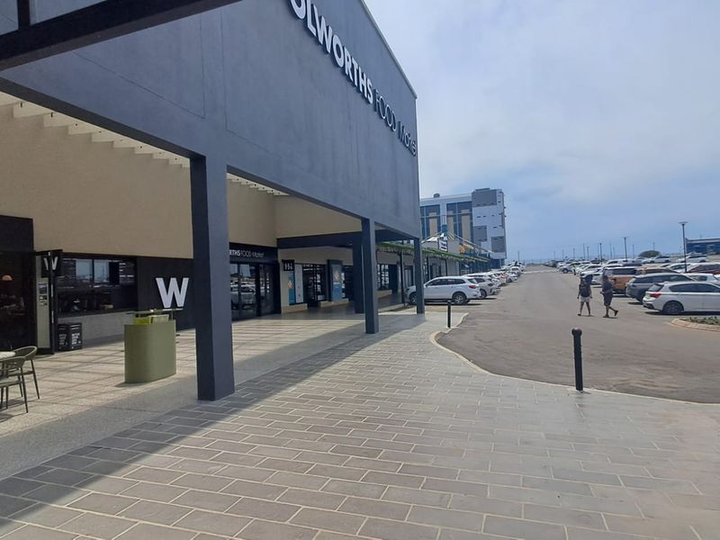 Exterior view of a modern retail storefront with a covered walkway, paved pedestrian area, and adjacent asphalt parking lot.