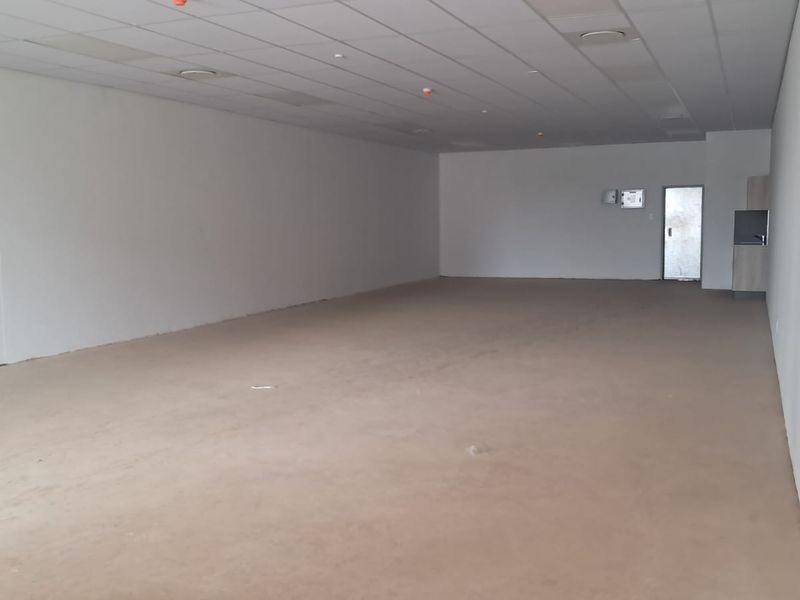 A spacious, empty open-plan retail unit featuring a suspended ceiling, recessed lighting, and a small kitchenette area.