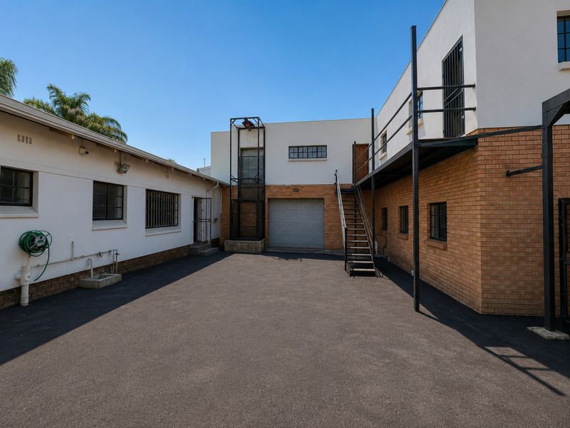 Rear exterior view of the industrial property featuring an asphalt courtyard, roller shutter door access, and an external metal staircase leading to an upper level.