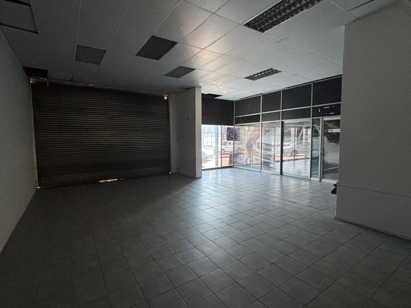 Vacant retail space featuring tiled flooring, a suspended ceiling with recessed lighting, a secure metal roller shutter, and a large glass storefront facade.