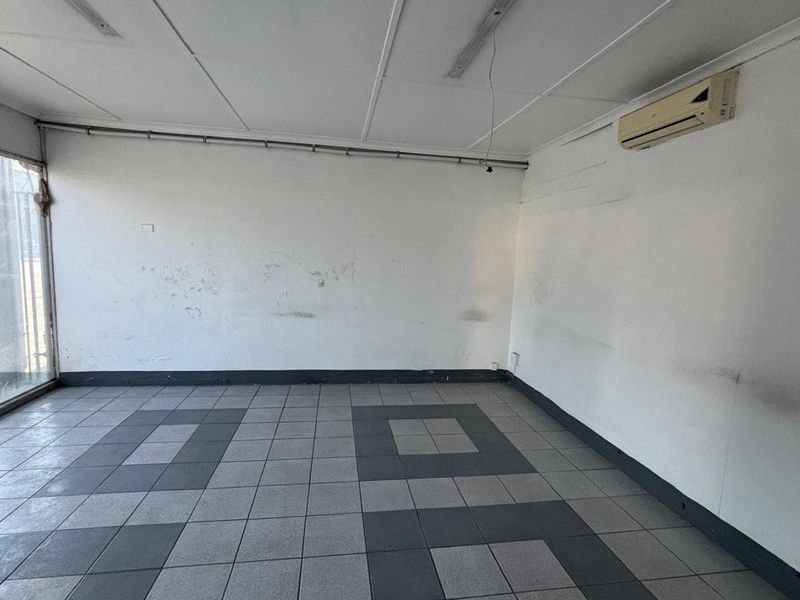 An empty retail space featuring tiled flooring, a suspended ceiling, and a wall-mounted air conditioning unit, requiring cosmetic updates to the wall finishes.