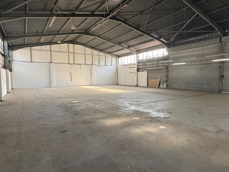 Spacious industrial warehouse unit featuring high ceilings with exposed steel trusses, concrete flooring, and a large roller shutter door for easy access.