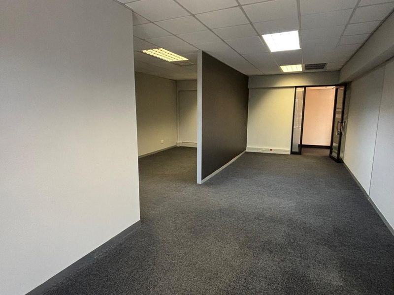 An empty, open-plan office space featuring neutral walls, carpeted flooring, a partition wall, and recessed lighting within a suspended ceiling.