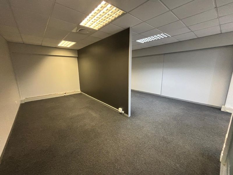 An empty office space featuring a central partition wall, suspended ceiling with recessed lighting, and grey carpet flooring.