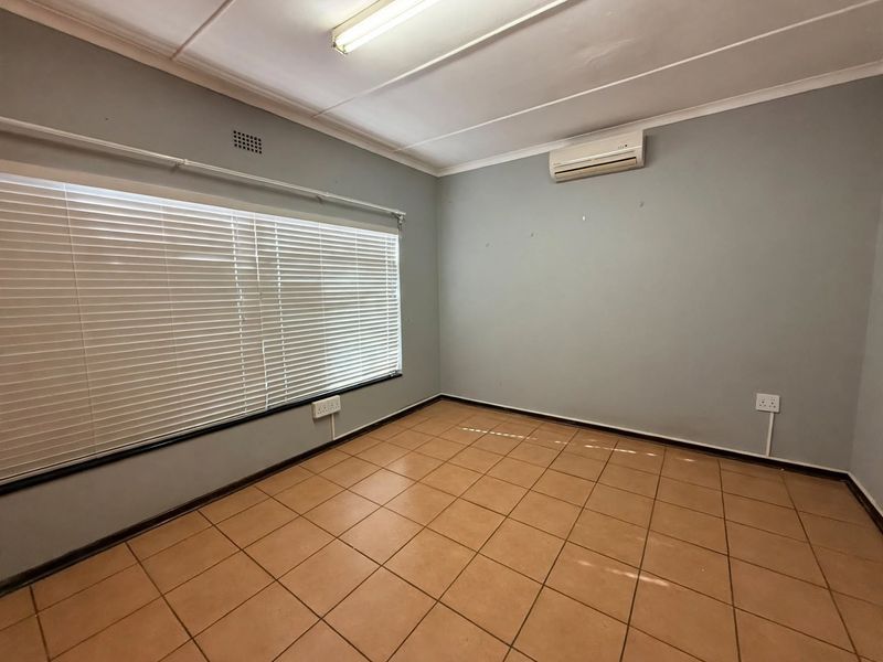 A clean, empty office space featuring tiled flooring, an air conditioning unit, and large windows with fitted venetian blinds.