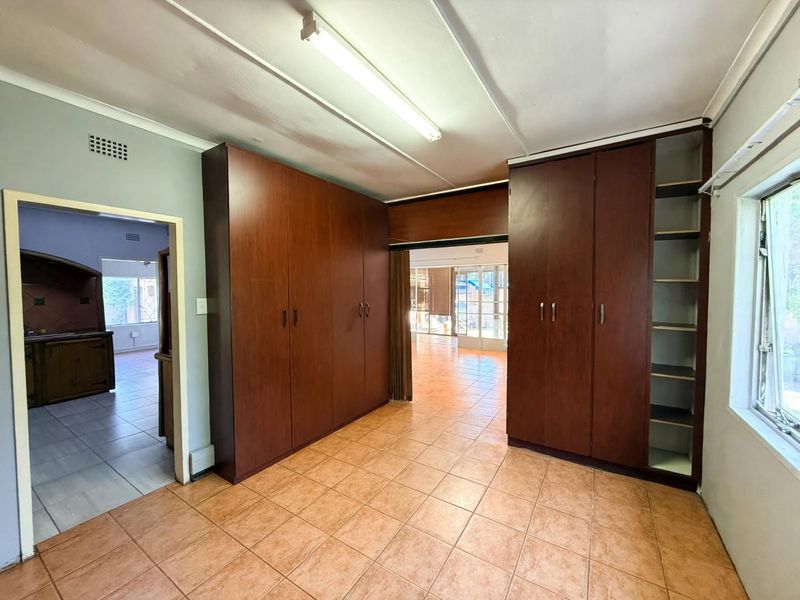 An empty office space featuring built-in wooden cupboards, tiled flooring, and fluorescent lighting, offering a functional layout with views into adjacent rooms.