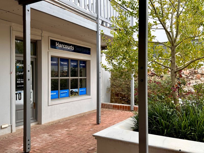 Front exterior view of the office premises featuring a brick-paved courtyard, stone retaining wall, and a covered walkway with an overhead balcony structure.