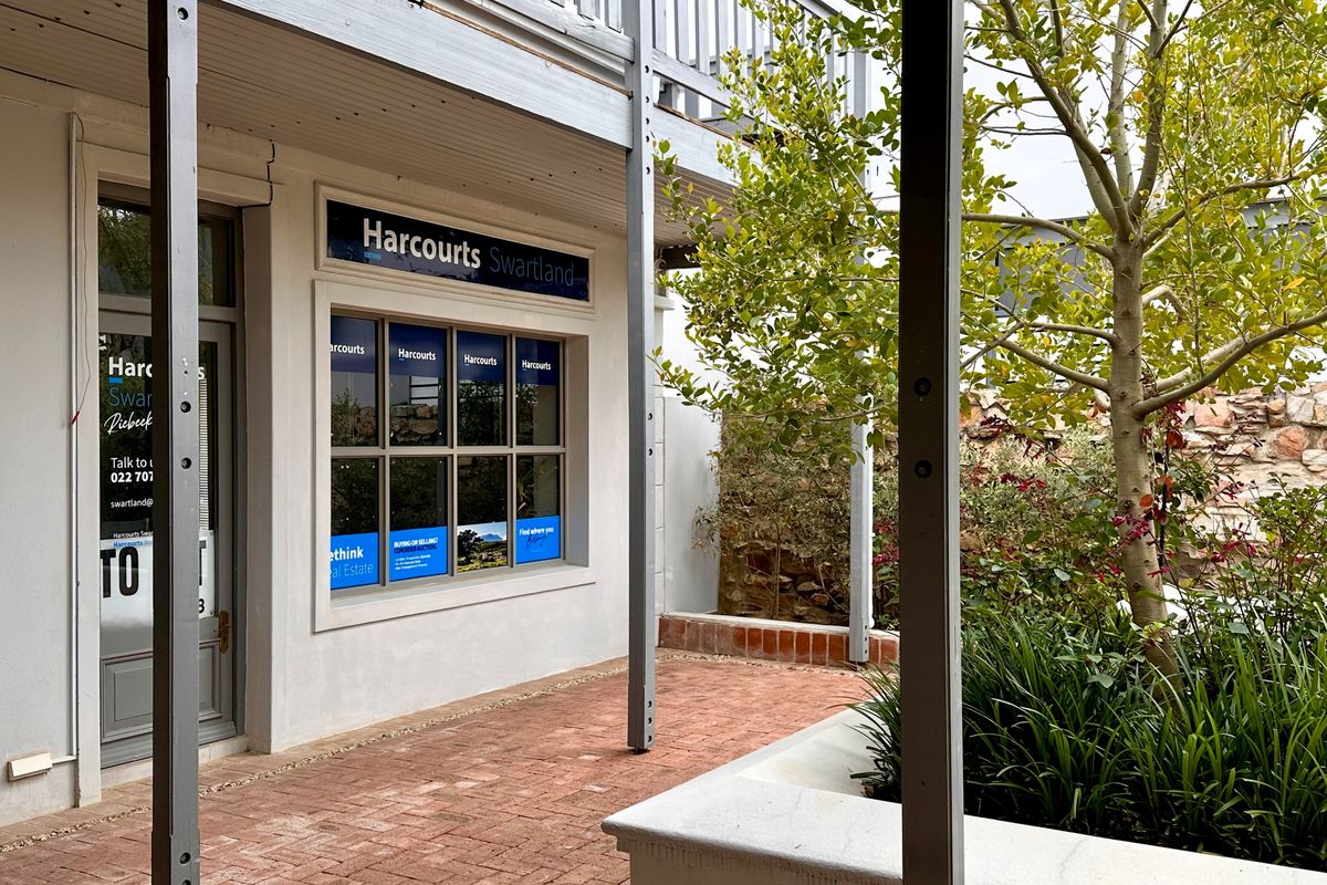 Front exterior view of the office premises featuring a brick-paved courtyard, stone retaining wall, and a covered walkway with an overhead balcony structure.