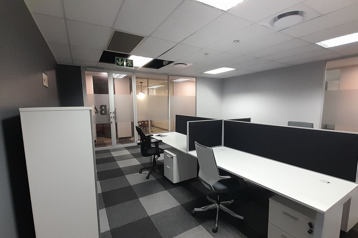 A functional office space featuring glass partitions, carpeted flooring, and built-in desk workstations with integrated privacy screens.
