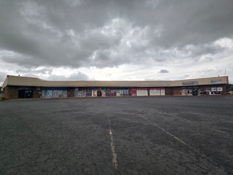 A wide-angle view of a commercial strip mall featuring multiple storefronts and a large, weathered asphalt parking lot under an overcast sky.