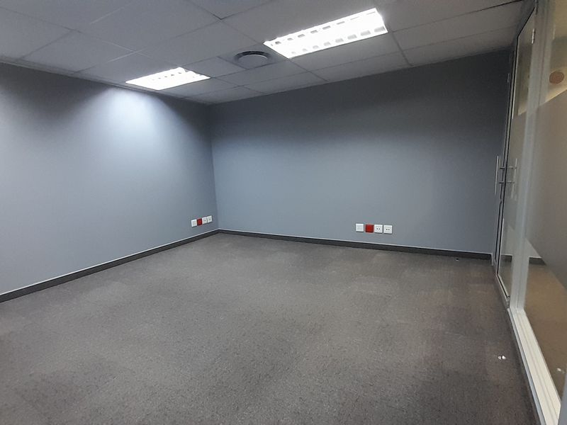 A clean, empty office space featuring grey walls, carpeted flooring, recessed ceiling lighting, and a modern glass partition wall.