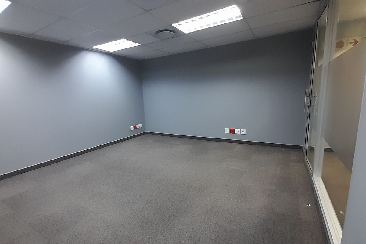 A clean, empty office space featuring grey walls, carpeted flooring, recessed ceiling lighting, and a modern glass partition wall.