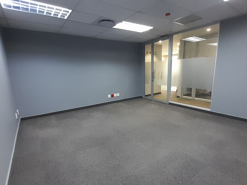 A clean, empty office space featuring grey carpet tiles, neutral walls, a glass partition wall, and recessed ceiling lighting.