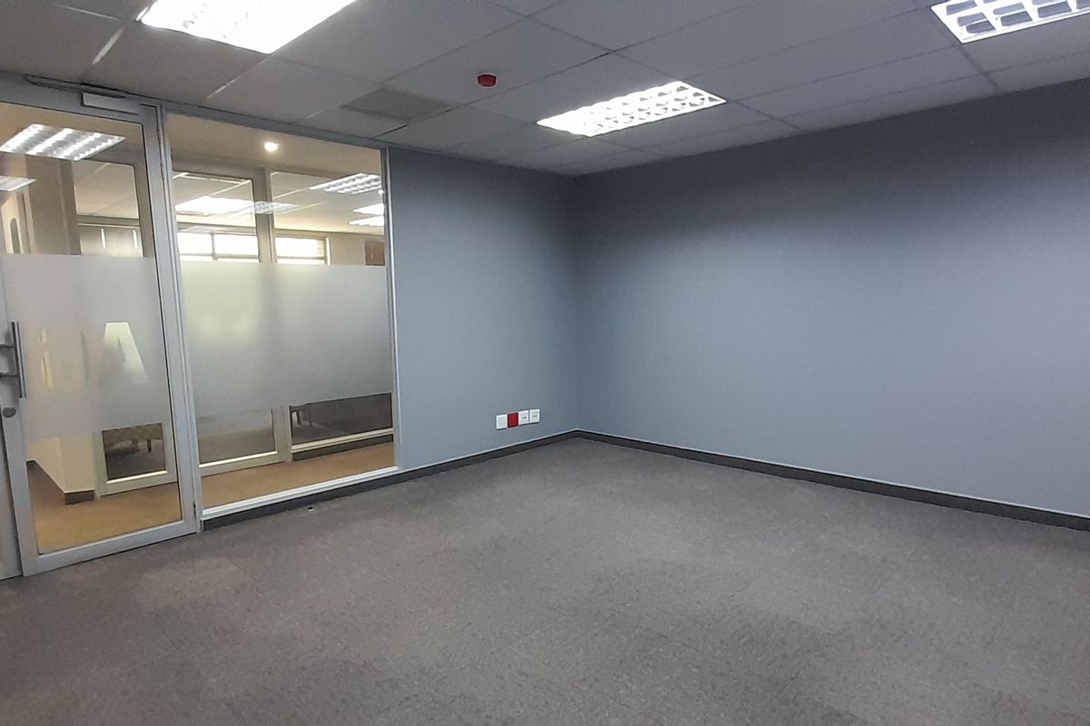 A clean, empty office space featuring grey walls, carpeted flooring, a glass partition wall, and recessed ceiling lighting.
