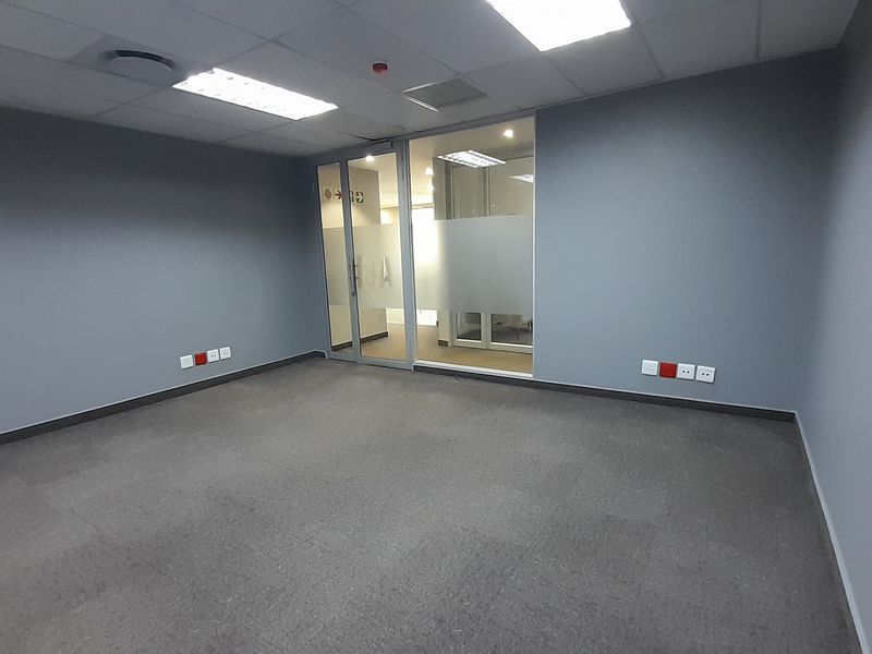A clean, vacant office space featuring grey carpet tiles, neutral walls, a glass partition entrance, and integrated ceiling lighting with power and data points.