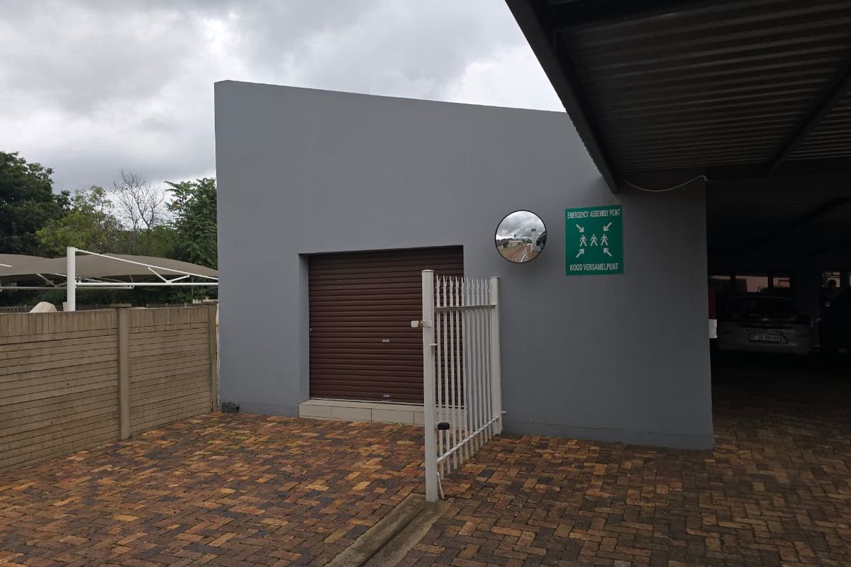 Exterior view of a commercial building featuring a paved courtyard, roller shutter door, metal security gate, and covered parking area.