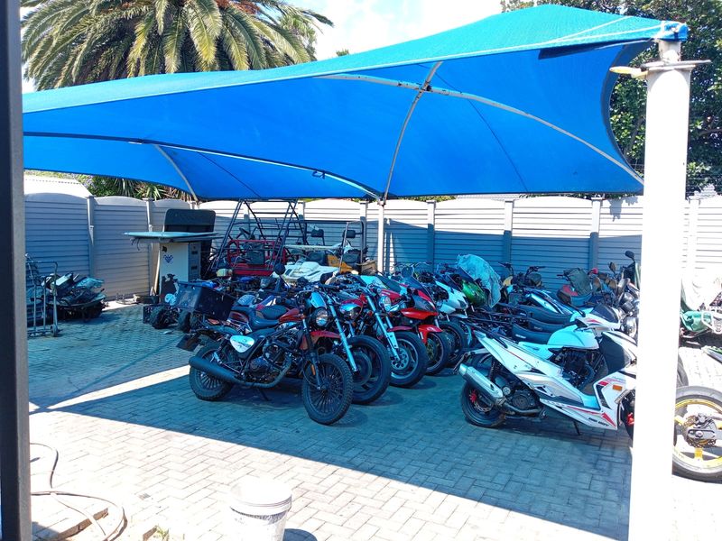 A paved outdoor area featuring a large blue shade sail structure, providing covered parking or storage space for multiple motorcycles.