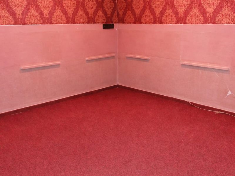 An empty commercial office space featuring red patterned wallpaper, carpeted flooring, wall-mounted shelving ledges, and an integrated air conditioning unit.