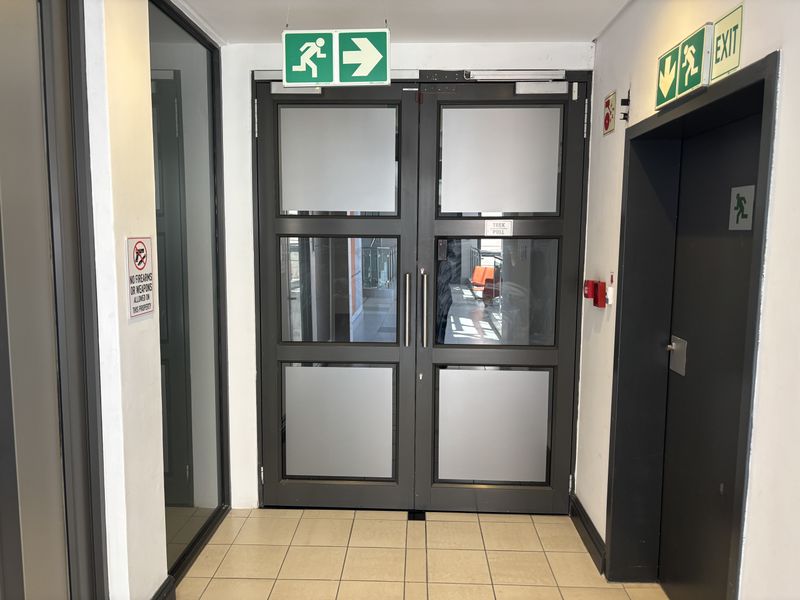 A clean, functional office hallway featuring double glass doors, fire safety signage, and durable tiled flooring.
