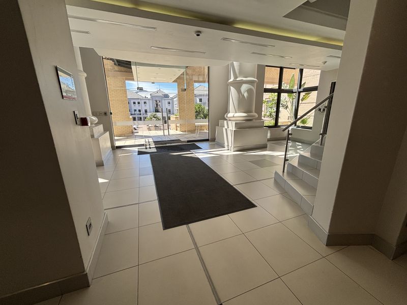 Bright and spacious office building entrance lobby featuring tiled flooring, a decorative pillar, and a staircase with modern stainless steel railings.