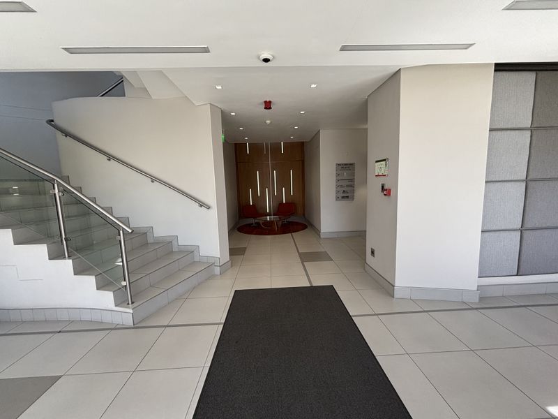 Modern office building entrance lobby featuring tiled flooring, a staircase with glass balustrades, and recessed ceiling lighting.