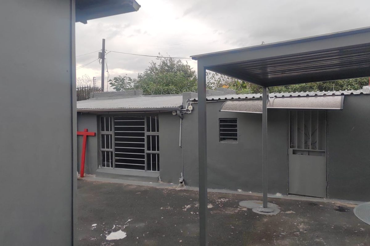 Exterior view of a commercial property featuring a paved courtyard area, covered patio structure, and secure metal window bars.