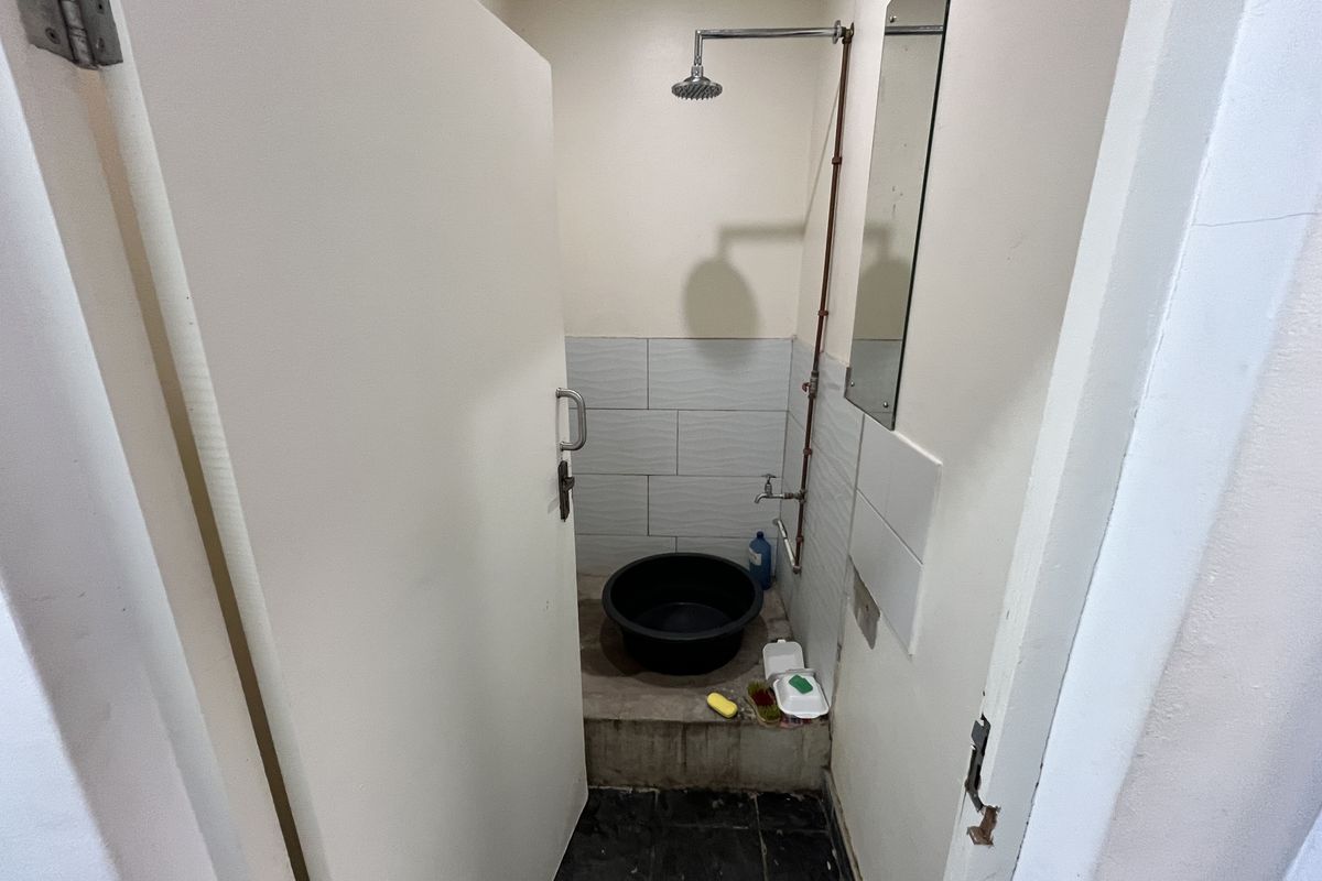 A small, basic bathroom facility featuring a wall-mounted shower head, exposed plumbing, and tiled splashback area.
