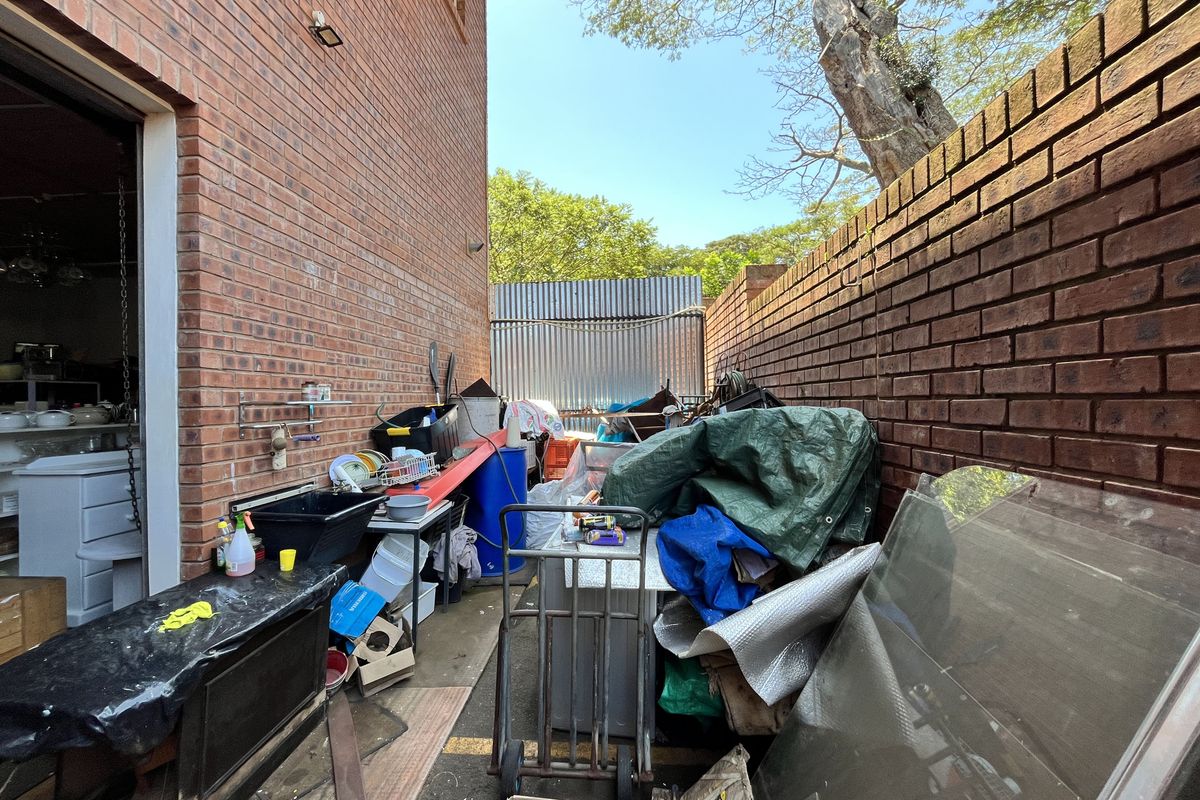 A narrow exterior side passage featuring exposed brick walls and a corrugated metal boundary fence, currently used for storage.