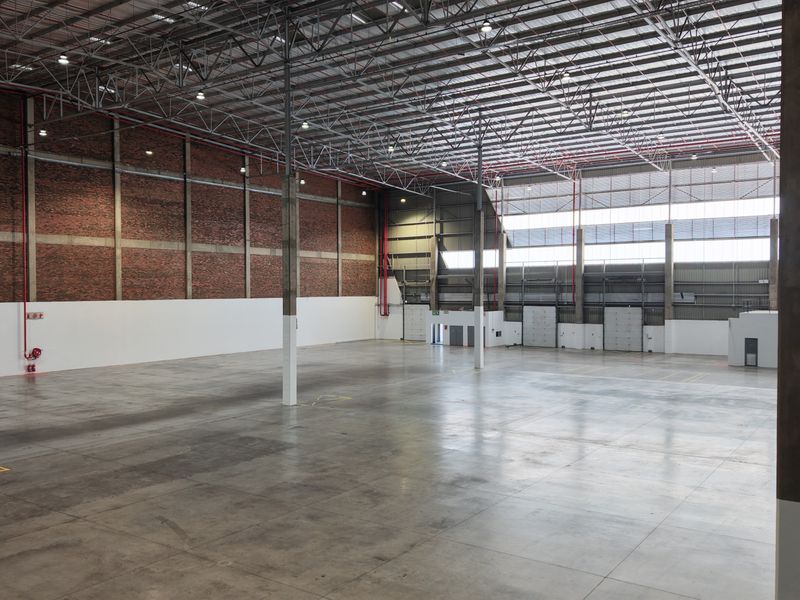 Spacious industrial warehouse featuring high ceilings with exposed steel trusses, concrete flooring, exposed brick walls, and multiple roller shutter doors for efficient logistics.