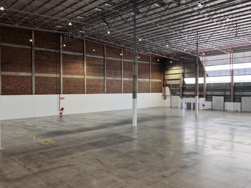 Spacious industrial warehouse featuring high ceilings with exposed steel trusses, concrete flooring, exposed brick walls, and multiple roller shutter doors.