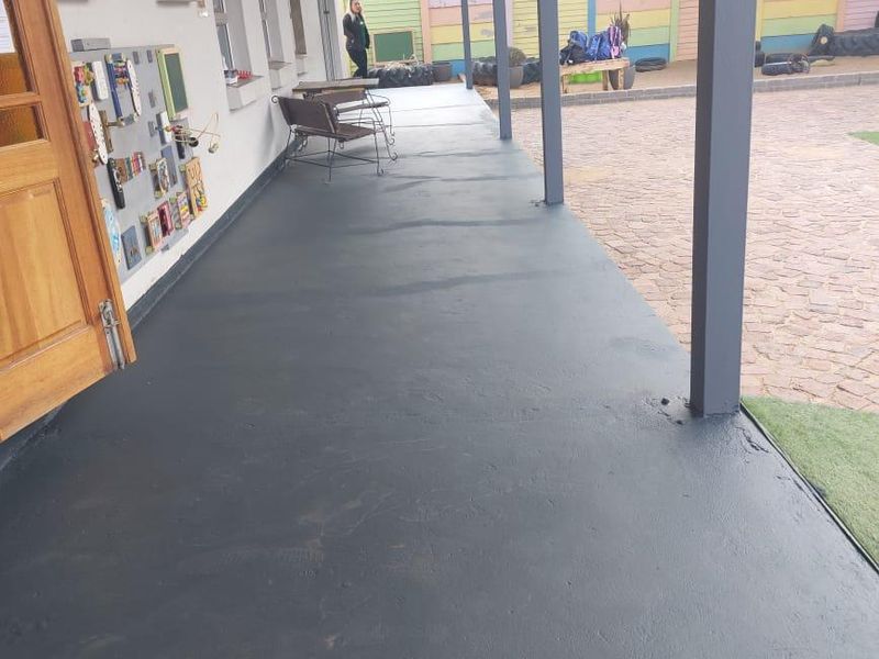 A covered outdoor patio area featuring painted concrete flooring, support pillars, and access to a paved courtyard space.