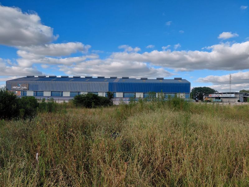View of a large commercial industrial warehouse building with blue metal cladding, situated on a vacant land plot with overgrown vegetation.